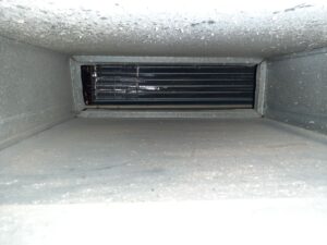 The image shows the inside of an air duct with gray metallic walls and a visible round black fan or vent at the far end. The surfaces appear dusty, indicating the duct may need cleaning. The photo is taken with a bright flash.