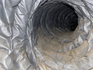 Close-up view inside a flexible, dirty air duct or ventilation hose, showing accumulated dust and debris on the inner ridges. The duct is gray with a spiral, ribbed texture, and the light highlights the grime along the surface.