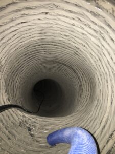 Close-up view inside a dusty, circular air duct with ridged metal walls. Two hoses, one black and one blue, extend inward, likely for cleaning or maintenance purposes. Light reflects off the dust-coated interior surfaces.