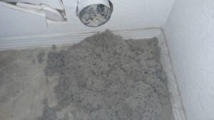 A large pile of gray lint is scattered on the floor in the corner of a room, directly below an open dryer vent duct protruding from the wall. The duct and wall are dirty, and lint buildup is evident. The floor is tiled.