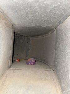 Inside a dusty metal air duct, a colorful ball of yarn and a small orange-tipped screwdriver rest on the floor. The surfaces are coated with dust and dirt, and the space is dimly lit.