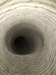 Close-up view inside a circular, flexible air duct or vent. The duct’s inner surface has ridged, spiraling layers, appearing metallic or plastic, and is illuminated by a flash, highlighting its reflective, textured interior, with darkness at the far end.