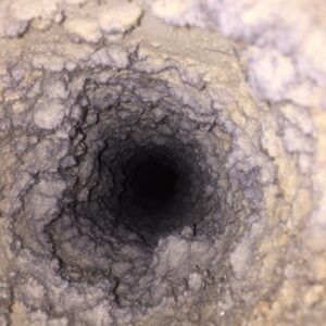 Close-up view inside a vent or duct, densely lined with thick, gray lint and dust buildup, forming a tunnel-like shape with a dark center. The texture is rough and fibrous, indicating significant accumulation of debris.