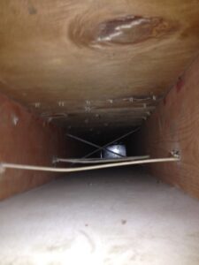 View inside a rectangular air duct with wooden walls and ceiling. The image shows some metal support wires and dust on the floor of the duct, with light reflecting at the far end. The scene appears enclosed and slightly out of focus.