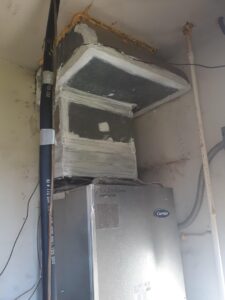 A silver Carrier HVAC unit is installed in a corner. Above it is a boxy duct covered in insulation, silver tape, and sealant. A thick black insulated pipe runs vertically along the left, and the walls show stains and signs of wear.