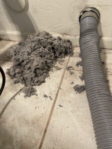 A pile of gray dryer lint is on a tiled floor next to a flexible gray dryer vent hose that is detached from the wall. The lint appears loose and messy, with bits scattered around the main pile. The wall behind shows a vent opening.