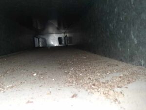 Interior view of a metal air duct, with the camera looking down the length of the duct. The floor is covered in dust and dirt particles, with light reflecting off the metal surfaces. The scene appears dimly lit except for a flash.
