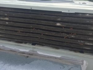 Close-up of an air conditioning unit with bent, dirty fins and visible debris caught in the coils. The metal slats are uneven and damaged, affecting airflow. Some leaves and dirt are lodged between the fins, and the surrounding surface shows signs of water stains.