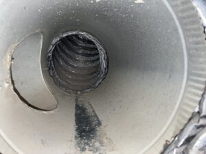 Close-up view inside a dusty metal air duct, showing a flexible black inner hose with spiral ridges. The duct interior has a buildup of dust and debris, and light shines from the open end, highlighting the dirt accumulated along the bottom.