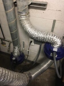 Two flexible silver ventilation ducts are attached to round blue machines on a concrete floor beside a white cinderblock wall. The ducts connect to vertical metal pipes. An electrical outlet with plugs and cords is visible on the wall.