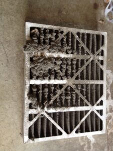 A dirty air filter lies on a concrete floor. The filter is covered in thick, grey dust and debris, especially concentrated in clumps along the left side. The accumulated dirt partially obscures the filter’s grid pattern.
